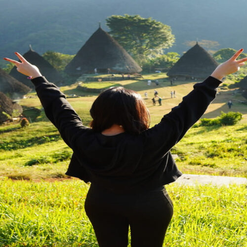 Open trip Wae Rebo: wisatawan mengangkat kedua tangan di depan rumah adat kerucut Mbaru Niang di tengah rumput hijau